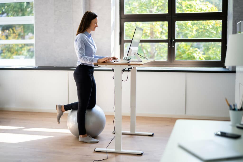 Höhenverstellbarer weißer Schreibtisch in Stehposition mit dunkelhaariger Frau und einem grauen Medizinball in hellem Office mit Fenstern ins Grüne