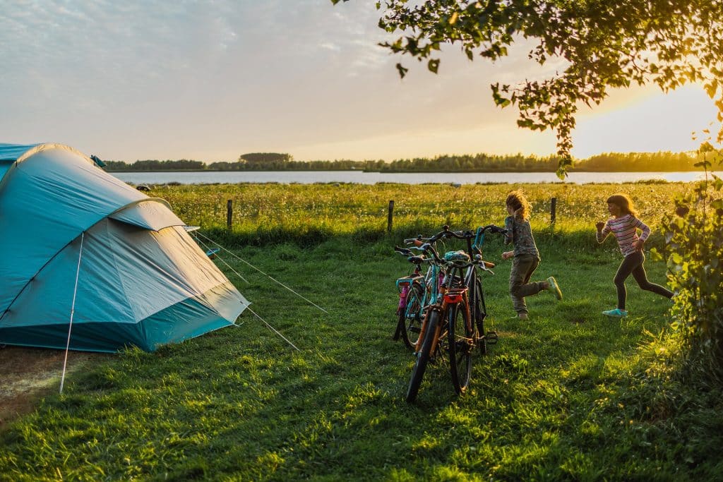 2 Kinder rennen fröhlich über eine Wiese im Sommer zu einem Campingzelt vor den Fahrräder stehen.