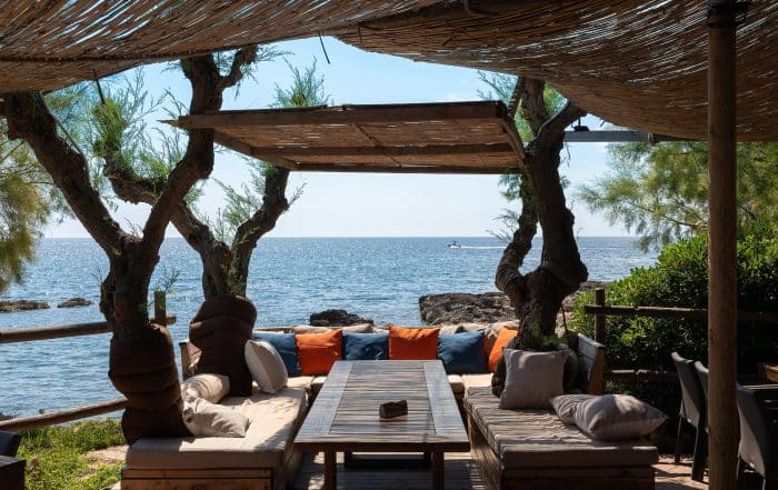 A table with comfortable benches to relax on the east coast of the Spanish island of Mallorca. There are many pillows on the cushions. In the background a boat is sailing across the water.