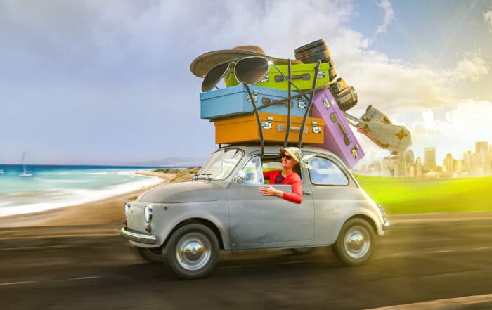 Fotomontage. Fiat 500 mit Frau am Steuer und übergroßen Koffern, Sonnenbrille, Baseballmütze und Fotoaparat auf dem Dach fährt auf einer Straße von Stadt im Hintergrund zum Strand