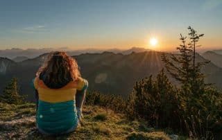 Wanderin sitzt im Graus auf einem Hügel und blickt in die untergehende Sonne übe den Bergen