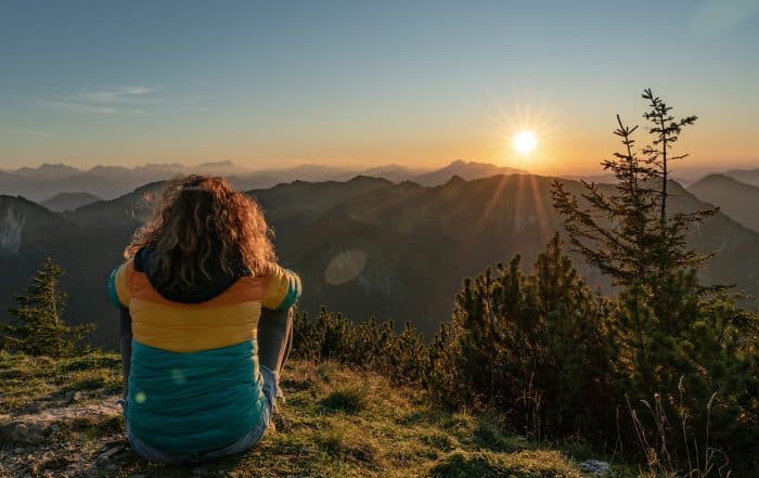Wanderin sitzt im Graus auf einem Hügel und blickt in die untergehende Sonne übe den Bergen