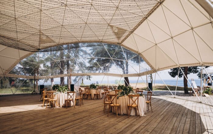 Großes kugelförmiges Gastronomiezelt an den Seiten offen mit Holzboden und runden Tischen mit weißen Tischdecken und Holzstühlen in einer Landschaft an der Küste