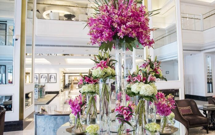 Runder Glastisch in einer modernen in weiß gehaltenen Hotellobby auf dem Glasvasen in verschiedenen Größen im Kreis angeordnet stehen in denen rosa Lilien und weiße Hortensienblüten stecken.