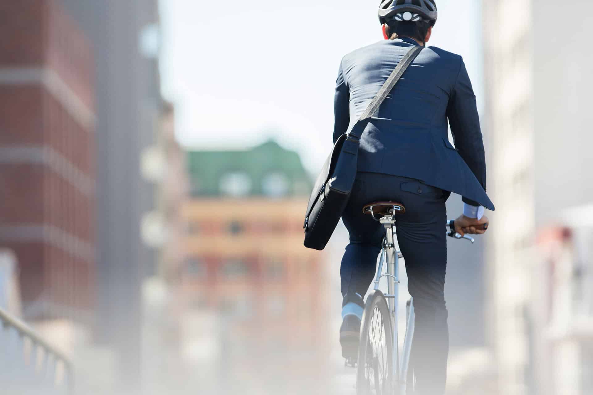 Junger Geschäftsmann mit Umhängetasche im Anzug sitzt lässig auf einem Fahrrad und fährt durch die Stadt. Aufnahme von hinten, Gebäude sind unscharf