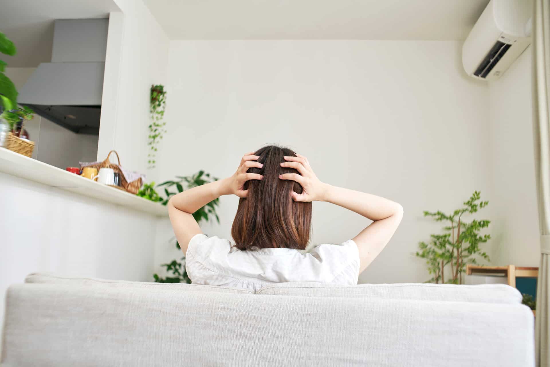 Gestresste Frau mit braunen Haaren sitzt auf dem Sofa und hält sich den Kopf mit beiden Händen. Fotografie zeigt die Frau von hinten