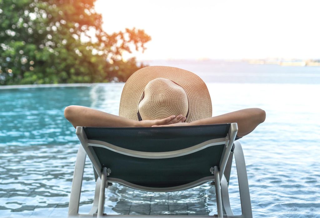 Frau mit Strohhut im Liegestuhl schaut über großen Pool aufs Meer