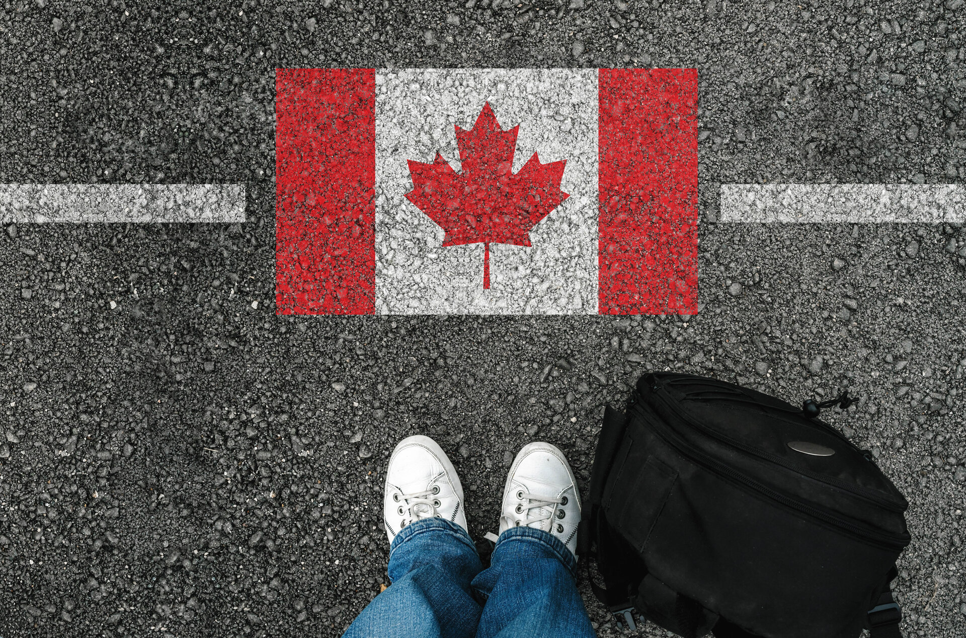 weiße Grenzlinie mit Kanadische Flagge auf einer Straße, davor eine schwarze Tasche und Beinpaar in Jeans und weißen Turnschuhen.