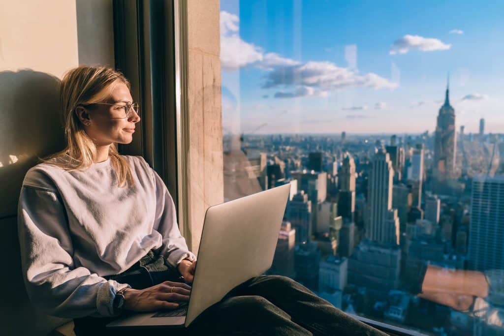 Junge Frau am Fenster mit Laptop blickt auf die Skyline New Yorks