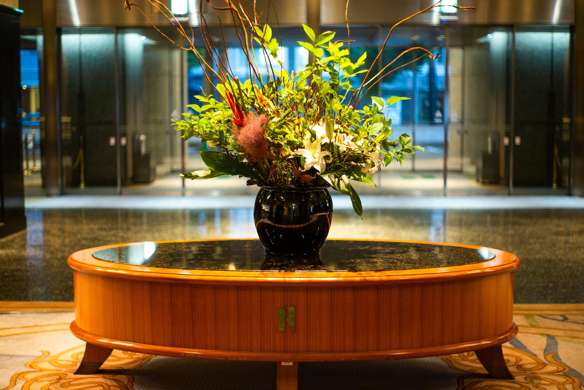 Hoteleingang mit niedrigem runden Holztisch mit Marmorplatte auf dem eine edle Vase mit geschmackvollem Blumenarrangement steht.