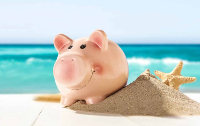 Rosa Sparschwein steht lächelnd auf einem Sandhaufen mit Seestern. Im Hintergrund unschaft türkisblaues Meer mit Wellen