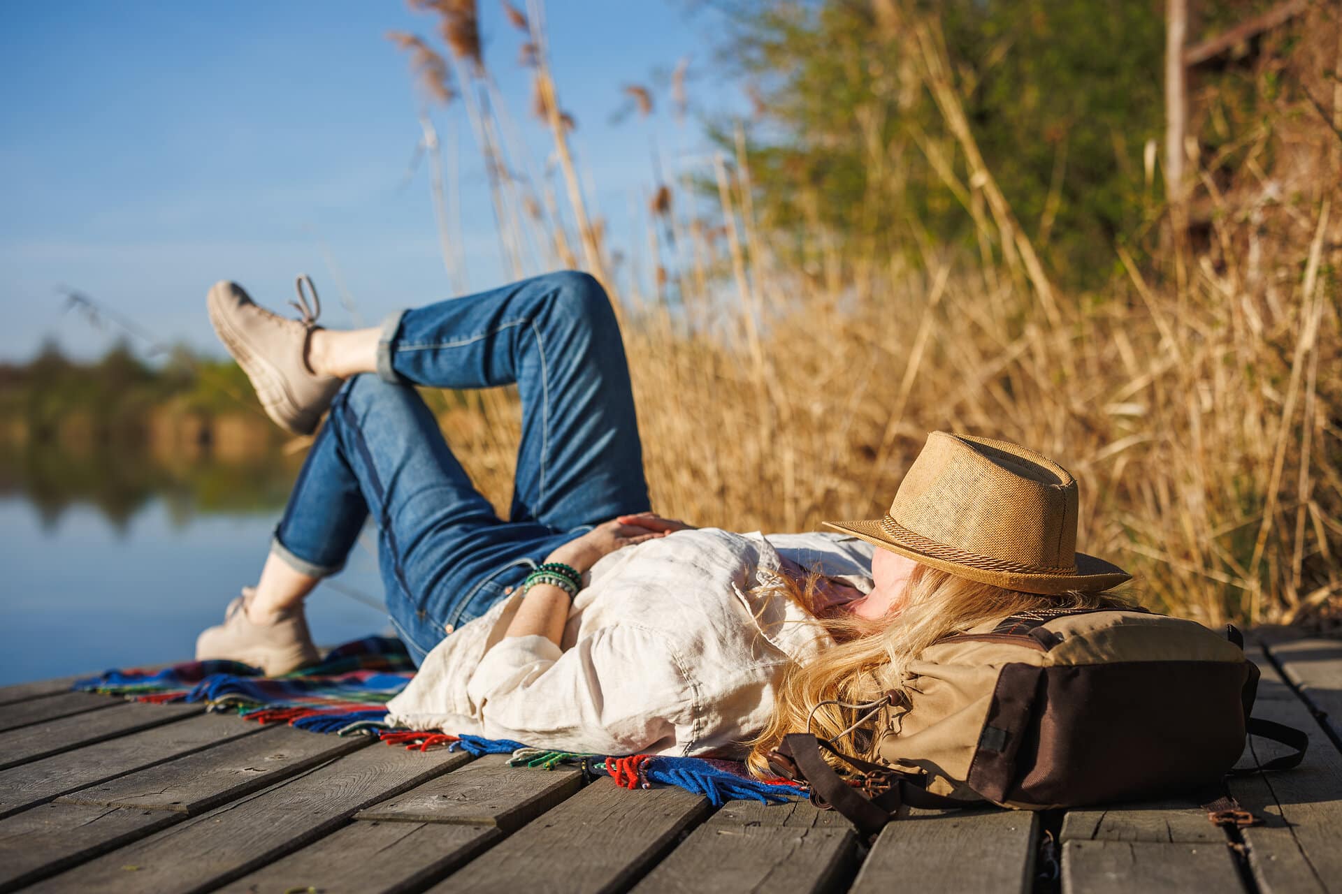 Frau liegt auf einem Holzsteg in der Sonne mit Strohhut auf dem Gesicht und mit dem Kopf auf eine Tasche und genießt den Moment der Ruhe