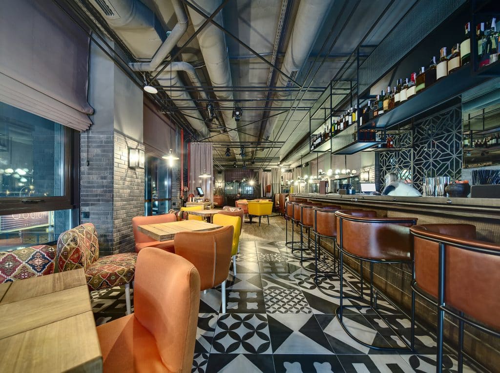 Glowing bar in a mexican restaurant in a loft style. On the right there is a bar rack with a laptop and accessories on it, brown chairs, shelves with dishes and accessories. On the left there are several wooden tables with multi-colored chairs, a brick wall with windows with curtains and glowing lamp, a red fire hose reel with red pipes, a wooden stand with a monitor and dishes. At the end there are another zones for seats. On the floor there are black and white tiles with patterns and a parquet. At the top there are lamps, speakers and communications. Horizontal.