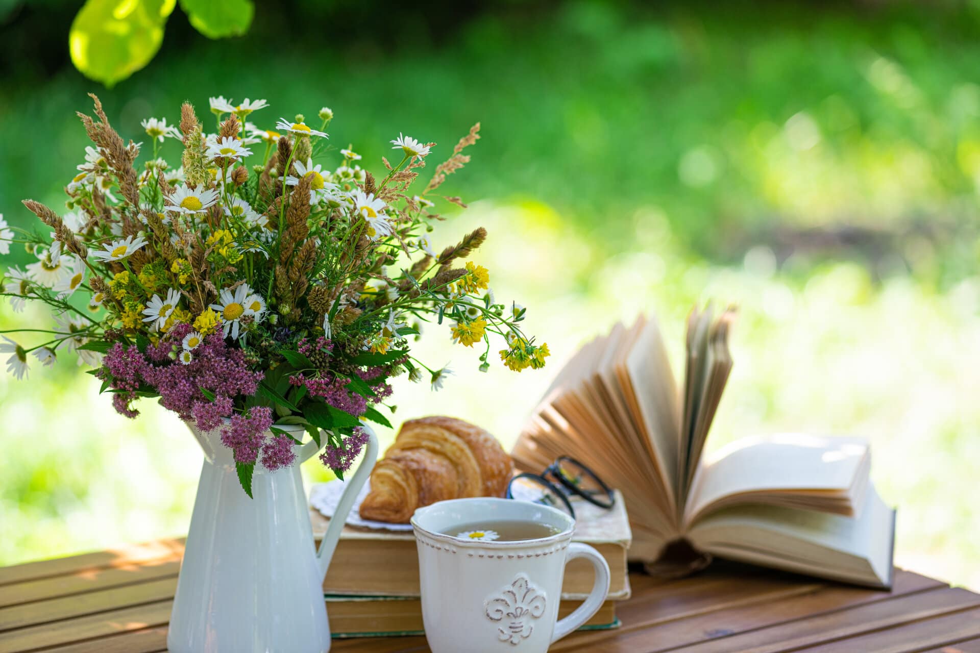 Wiesenblumenstrauß in weißer Porzellanvase auf Holztisch, daneben ein Teller mit Croissant eine Tasse Tee mit Gänseblümchen und ein aufgeschlagenes Buch, im Hintergrund sonniger grüner Garten