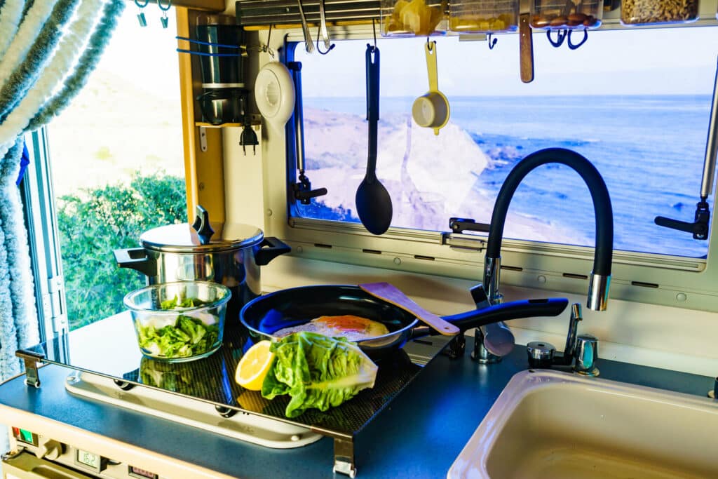 Ausblick von der Küche eines Campingmobils auf die Küste und das Meer auf dem Herd ist eine Pfanne mit Spiegeleiern und auf der Ablage liegen grüner Salat und eine Zitrone