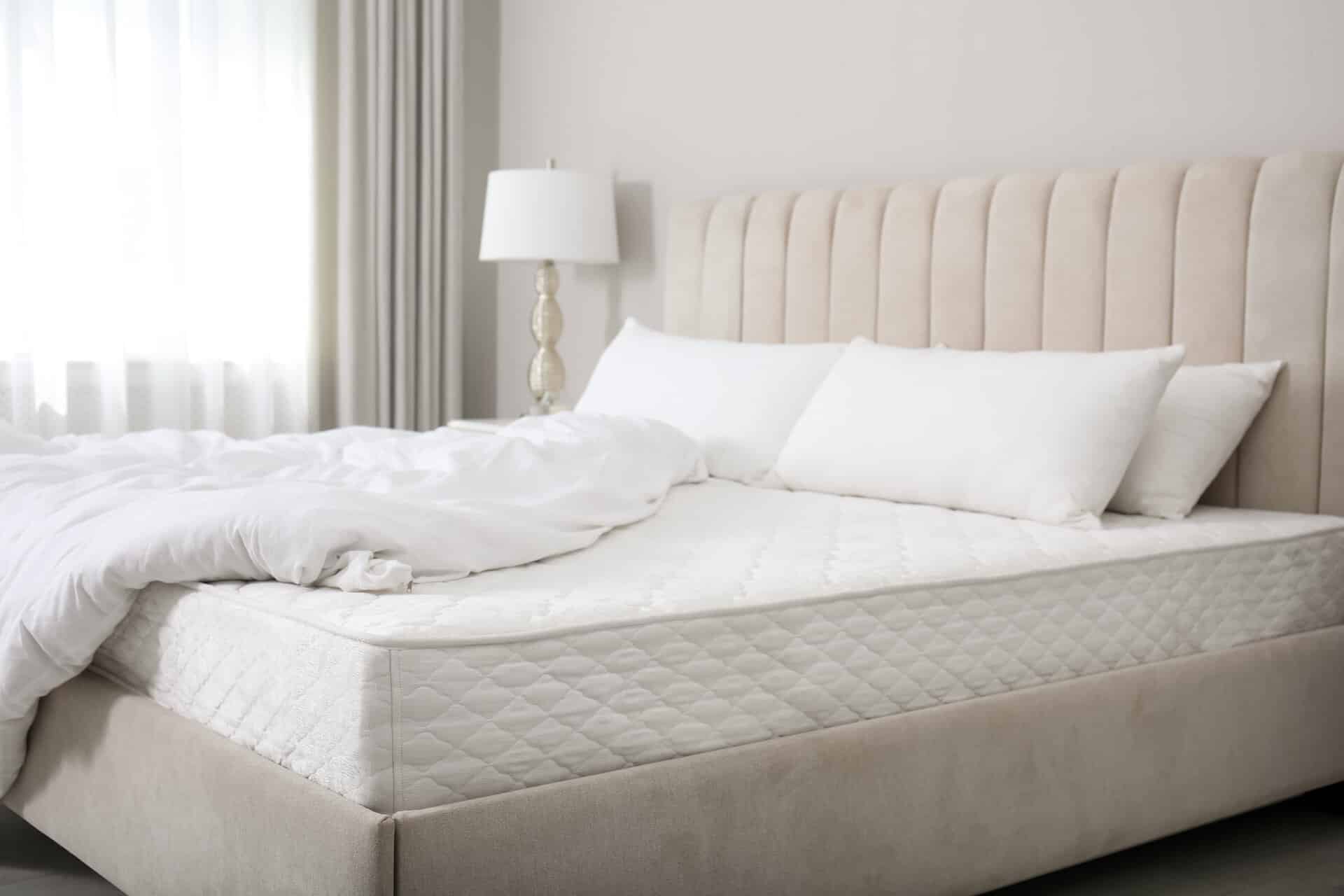 Komfortables Hotelbett in hellgrauem Tönen im hellen Zimmer mit weißen Kissen und weißer Bettdecke, die halb aufgedeckt ist.