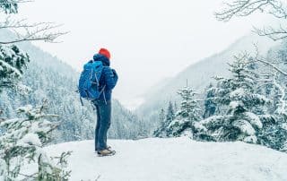 Wanderer mit Rucksack und warmer Kleidung blickt bei Schneefall in die verschneite hügelige und bewaldete Landschaft