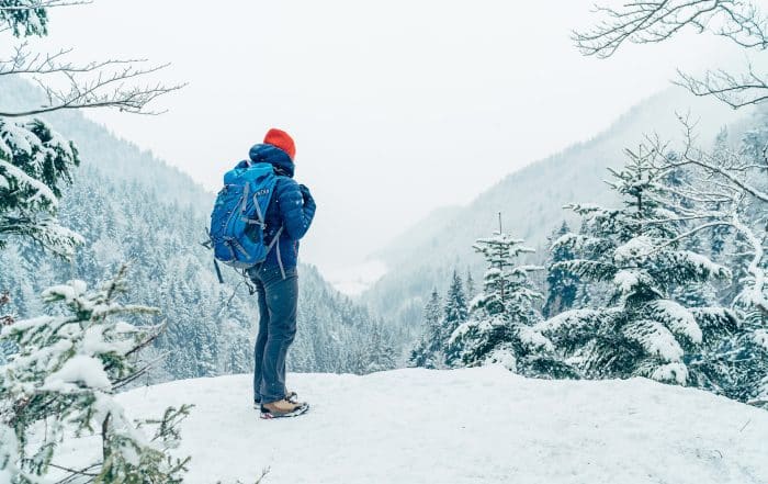 Wanderer mit Rucksack und warmer Kleidung blickt bei Schneefall in die verschneite hügelige und bewaldete Landschaft