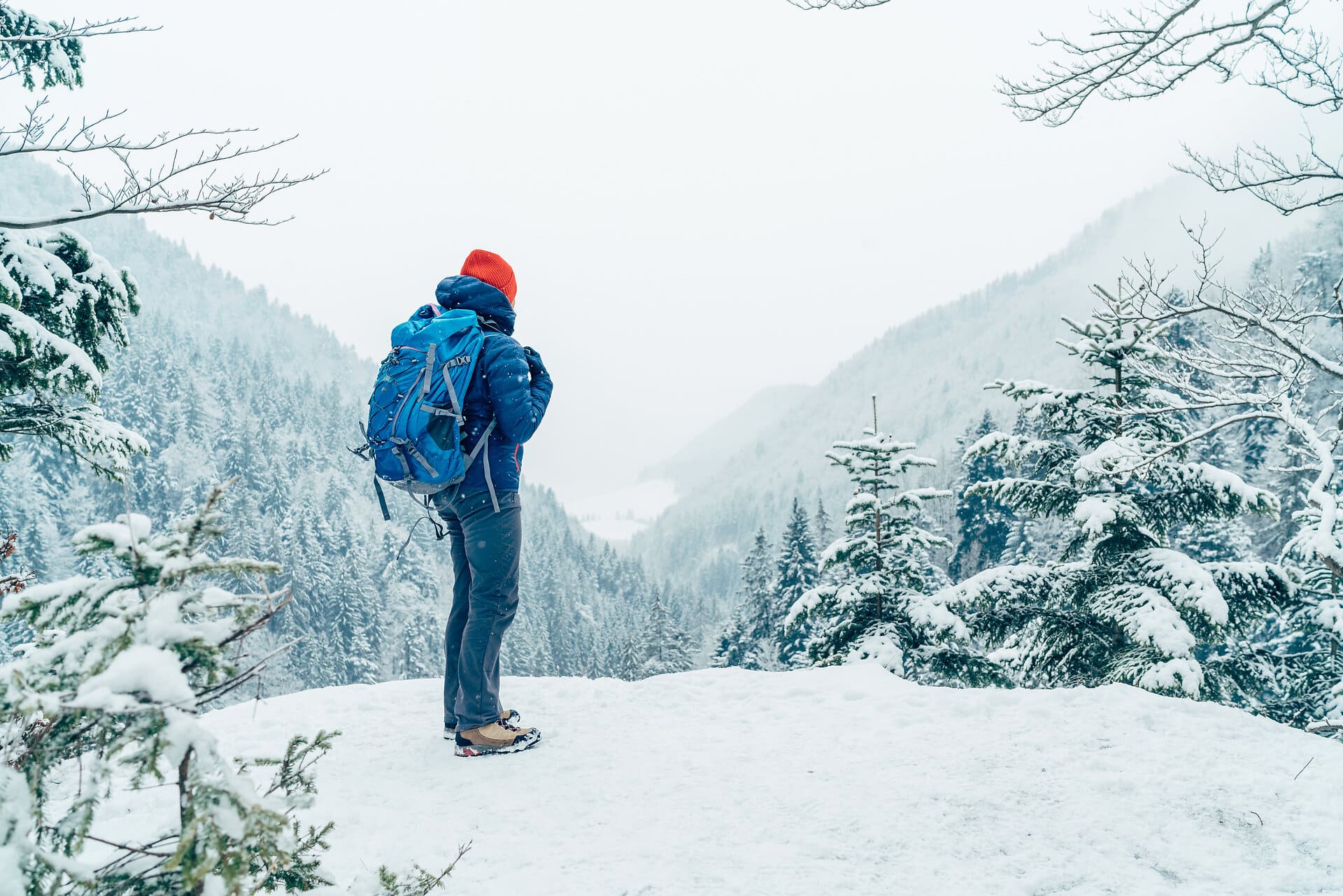 Wanderer mit Rucksack und warmer Kleidung blickt bei Schneefall in die verschneite hügelige und bewaldete Landschaft