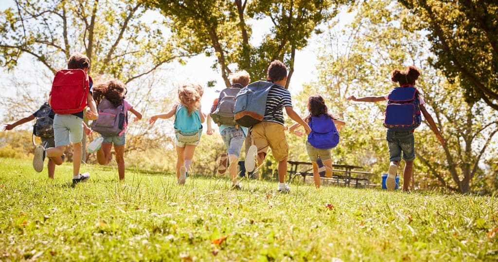 Grundschulkinder mit Rucksäcken im Sommer rennen über eine Wiese auf einen Rastplatz mit Holzbänken und Tischen zu