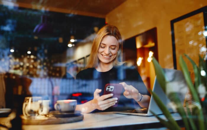 Junge Frau im Cafe mit Mobiltelefon und Notebook durch die Scheibe fotografiert.