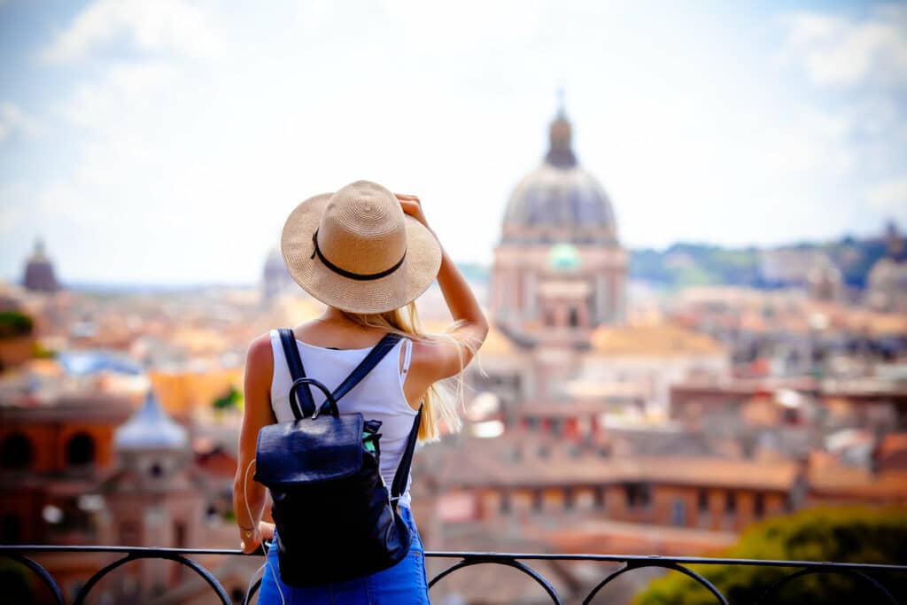 Junge blonde Frau mit Rucksack steht auf einem Aussichtspunkt in Rom und blickt auf die Stadt und den Vatican.
