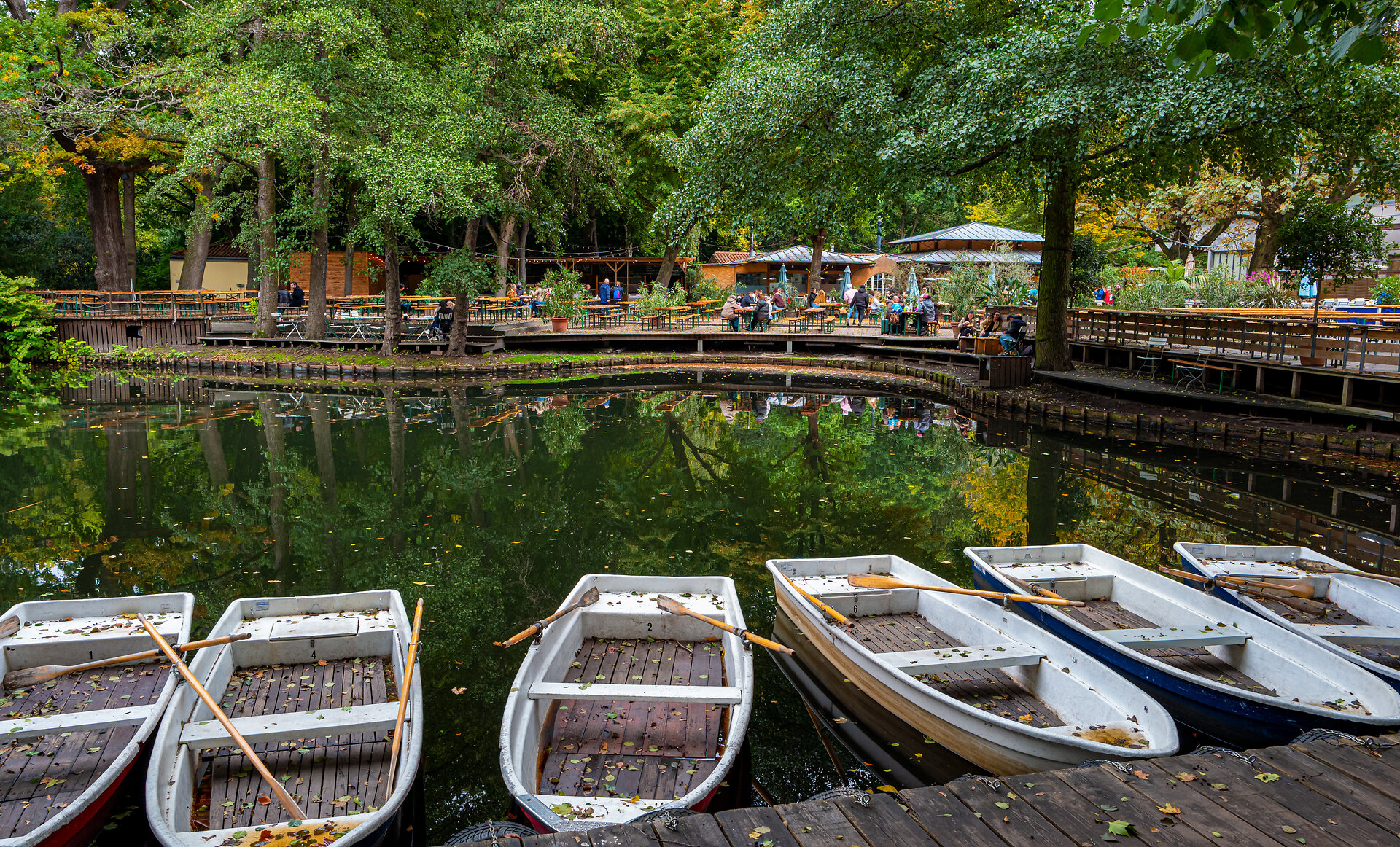 Ruderboote am Neuen See in Berlin im Herbst mit fallenden Blättern in den Booten im Hintergrund Biergarten mit Gästen