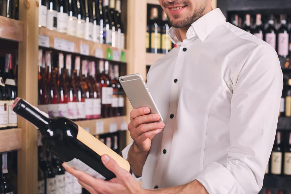 Junger Mann in Weinhandlung fotografiert das Etikett einer Weinflasche in seiner Hand