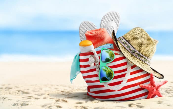 rot weiß gestreifte Badetasche am Sandstrand mit Meer im Hintergrund enthält Sonnenbrille, Flip-Flops, Sonnencreme, Handtücher und Strohhut