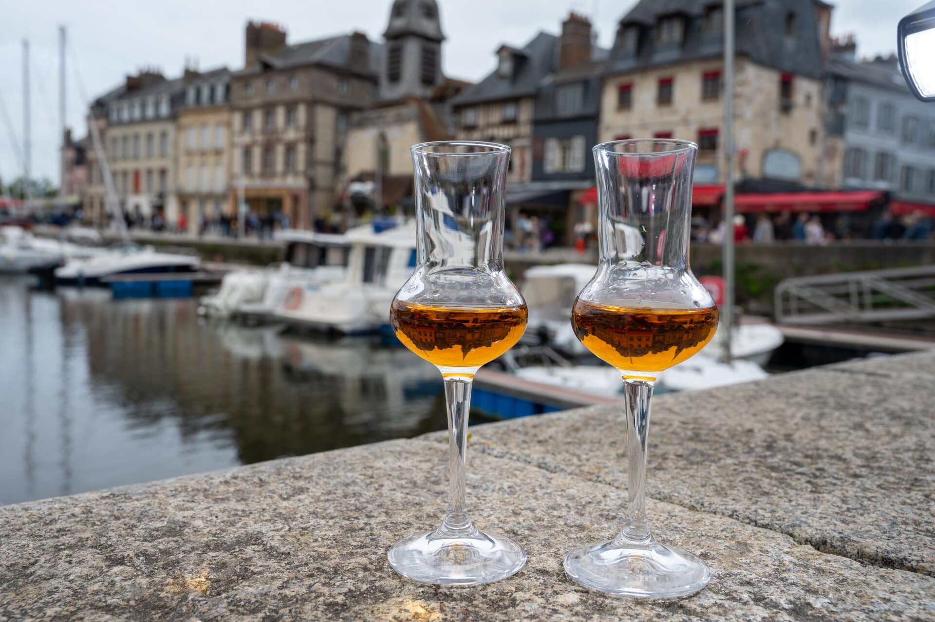 2 edle Gläser mit Calvados auf einer Kaimauer in der Normandie. Im Hingergrund Boote im Hafen und Häuserzeile.