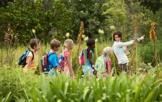 Lehrerin läuft mit einer Gruppe Schulkinder mit Rucksäcken durch einen Park und zeigt mit dem Arm nach vorne
