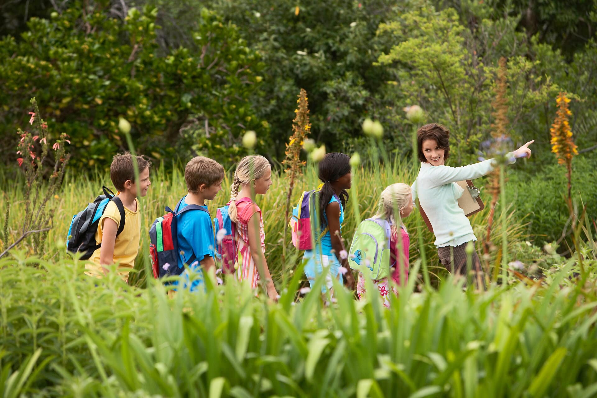 Lehrerin läuft mit einer Gruppe Schulkinder mit Rucksäcken durch einen Park und zeigt mit dem Arm nach vorne