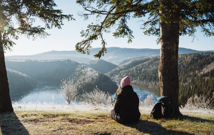Junge Frau sitzt auf einer mit Rauhreif überzogenen Wiese unter einer Fichte auf einem Hügel und blickt ins Tal und in die schöne Landschaft.