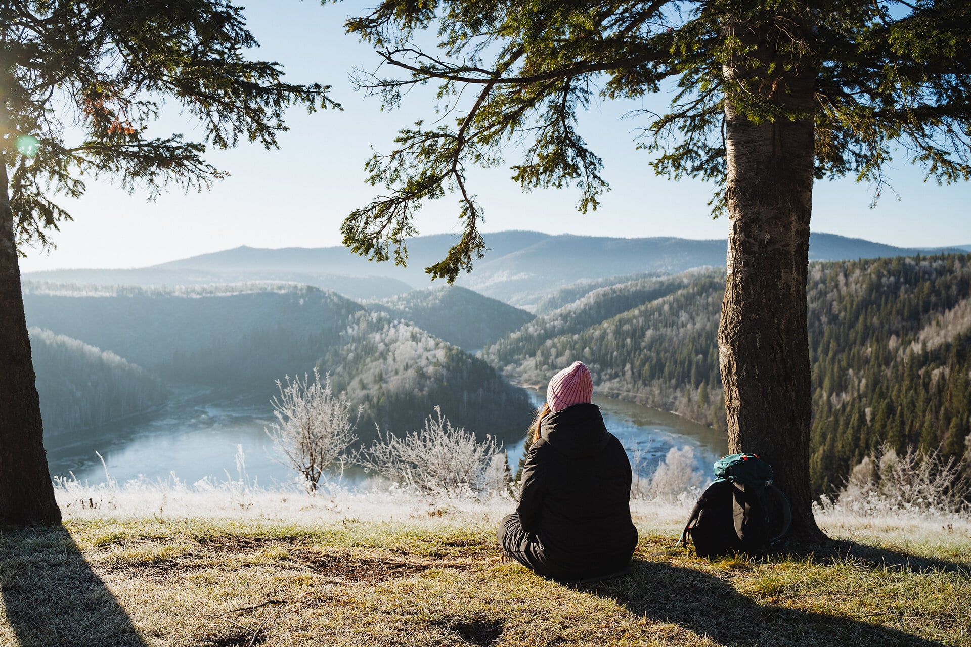 Junge Frau sitzt auf einer mit Rauhreif überzogenen Wiese unter einer Fichte auf einem Hügel und blickt ins Tal und in die schöne Landschaft.