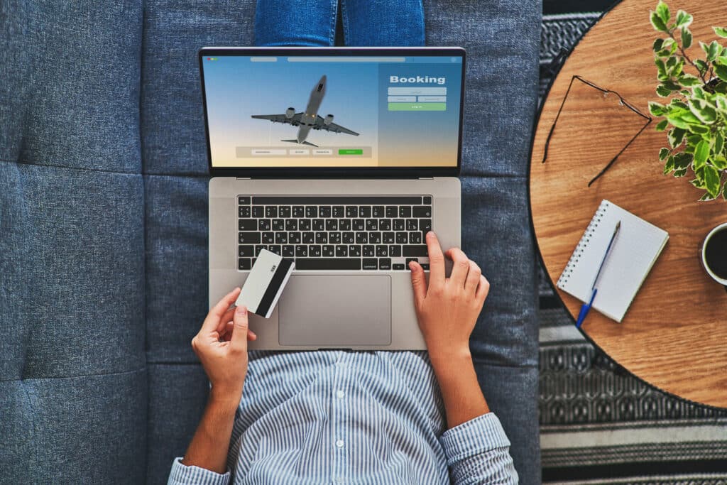 Ausschnitt einer Frau mit Notebook auf dem Schoß, die auf einer Couch liegt und eine Kreditkarte in der Hand hält. Auf dem Display ist ein Flugzeug und eine Buchungsmaske.