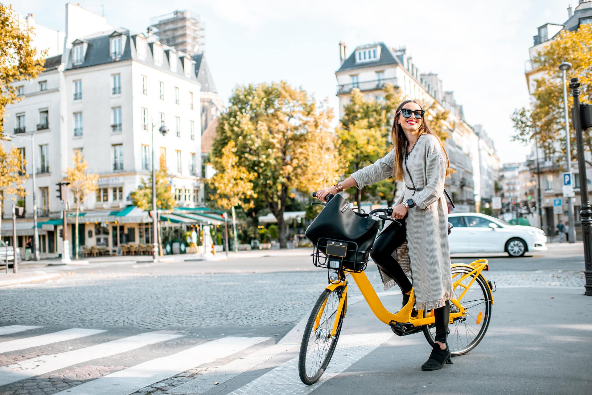 Junge Frau in modischer Kleidung und mit großer Sonnenbrille und schwarzer Ledertasche lächelt und steht mit schickem gelben Fahrrad an einer Kreuzung in Paris