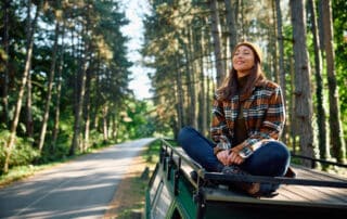 Junge Frau sitzt auf dem Dach eines Campers auf einer kleinen Straße im Wald und genießt den Moment mit geschlossenen Augen