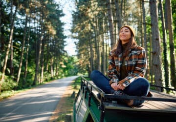 Junge Frau sitzt auf dem Dach eines Campers auf einer kleinen Straße im Wald und genießt den Moment mit geschlossenen Augen