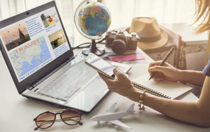 Junge Frau sitzt vor dem Laptop am Schreibtisch und plant eine Reise mit Handy und Reisewebsites und macht sich Notizen auf einem Block.