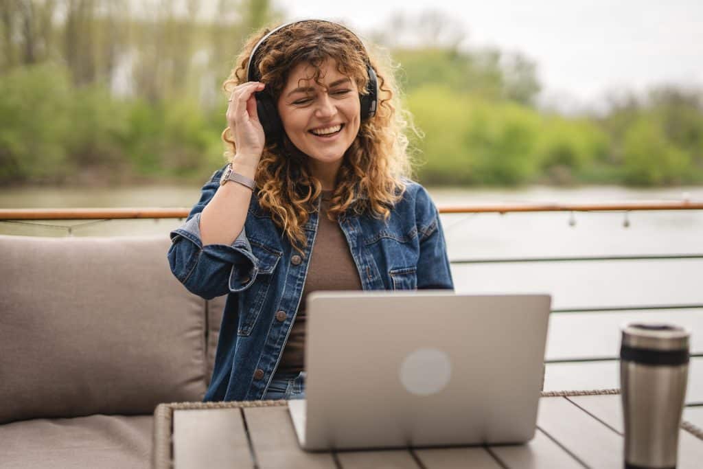 smiling adult woman wear headphones and sit on the terrace and have video call meeting on laptop