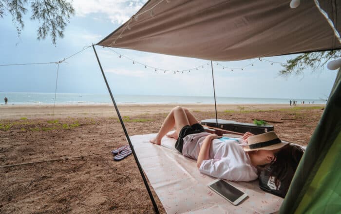 Frau liegt unter einem Sonnensegel auf einer Strandmatte mit Strohhut auf dem Gesicht in Shorts und weißem Hemd.