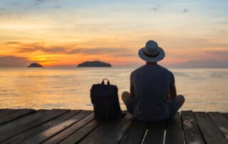Junger Mann mit Shorts und Sonnenhut sitzt auf einem Holzsteg und blick auf den Sonnenuntergang am Meer. Neben sich ein Rucksack