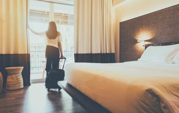 dunkelhaarige Frau mit Koffer im Hotelzimmer schaut aus dem Fenster - Sicherheitstipps
